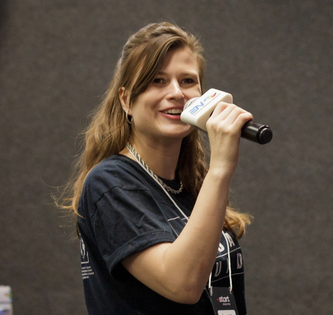 Foto de Débora Z. do Prado, cabelos longos loiros e segurando um microfone, simbolizando a expressão e a comunicação em um ambiente de apresentação.